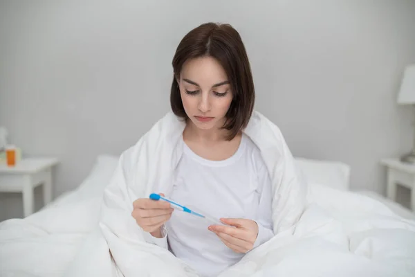 Young sick brunette woman with fever checking her body temperature with electronic thermometer, sitting in bed at home, lady got cold, flu or coronavirus, copy space