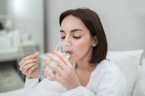 Unwell upset young brunette pretty lady sitting on bed, holding glass of water and pink capsule, sick woman taking painkiller at home, suffering from headache, period cramp, flu, closeup, copy space