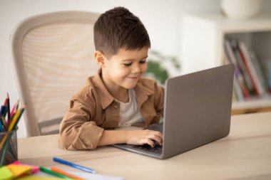 Video call remotely, education game. Glad caucasian little child pupil sits at table, typing on computer, play, watch video in room interior. Study at school, kindergarten and home, childhood and app