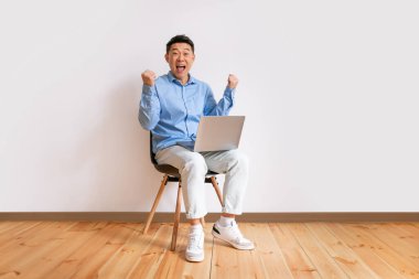 Happy asian businessman sitting on chair with laptop computer, raising hands up, excited over huge lottery win or business success, white studio wall, copy space, full length
