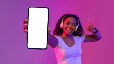 Mobile Offer. Cheerful Black Woman Showing Blank Smartphone With White Screen And Gesturing Thumb, Smiling African Lady Recommending New App, Standing In Neon Light Over Purple Background, Mockup