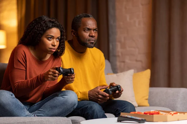 Concentrated funny black couple playing video games together in the evening in living room, man and woman sitting on couch, eating pizza, holding joysticks, looking at copy space. Home entertainment