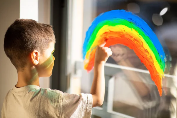 Caucasian little child draws rainbow with paint on window in elementary school, kindergarten room interior, close up. Fun alone, entertainment and play at home, art, education, fantasy and childhood
