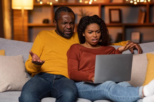 Furious shocked loving black couple mature husband and wife sitting on couch in cozy living room, looking at laptop screen, gesturing, reading news online, copy space