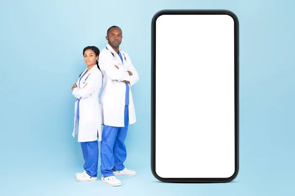 Black male and female doctors in white coats posing near big smartphone with blank screen isolated on blue background, mockup. Great offer, ad and app for remote health care and treatment