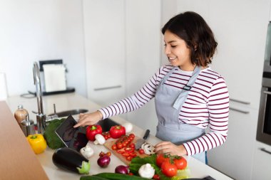 Side view of pretty young hispanic woman cooking delicious healthy food at home, using digital pad with black empty screen, checking nice cooking app on tablet, watching food vlog, copy space, mockup
