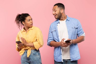 Black man with cellphone spying on his girlfriend chatting on mobile phone, posing on pink studio background. Couple using smartphones, man does not trust his woman