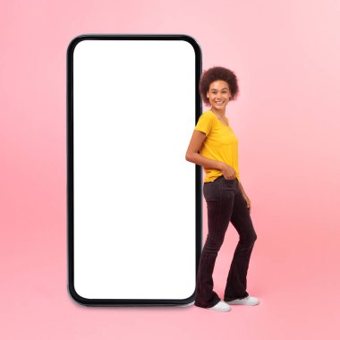 Smiling millennial multiethnic woman in casual stands near huge smartphone with empty screen isolated on pink background, studio. Blog, chat, website, app recommendation for work, study and lifestyle