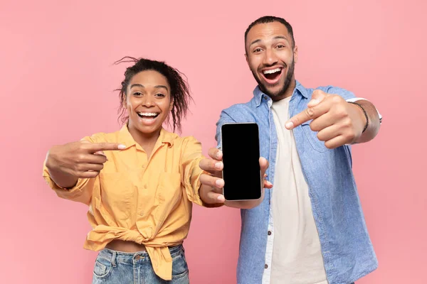 Emotional black couple pointing fingers at black smartphone screen, showing nice application or website on mobile phone, pink studio background, mockup