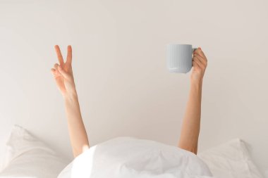 Young european woman lies on white bed raises her hands from under blanket holds cup of coffee and shows peace sign in bedroom interior, cropped. Good morning, vitality, weekend and wake up at home