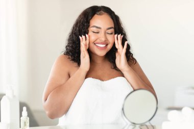 Facelift, beauty secrets concept. Happy plus size woman massaging her face, sitting in front of the mirror and smiling. Bodypositive lady doing anti wrinkle facial massage for skincare