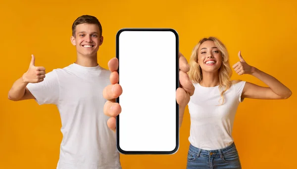 手に大きな空白のスマートフォンを保持し ジェスチャーを親指を表示笑顔カップル 幸せな男と女は新しいモバイルアプリケーションやウェブサイトをお勧めします 黄色の背景に立って モックアップ — ストック写真