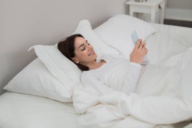 Happy pretty brunette young lady lying alone in big comfy bed at home, using cell phone and smiling, woman waking up with morning message from lover, side view, copy space