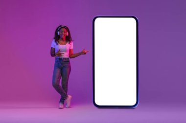 Mobile App. Happy Black Female Wearing Headphones Pointing At Big Blank Smartphone With White Screen, Cheerful African American Woman Standing In Neon Light Over Gradient Background, Collage, Mockup