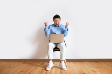 Overjoyed middle aged asian man with laptop computer sitting on chair, celebrating great deal or business success, making YES gesture against white studio wall, copy space, full length