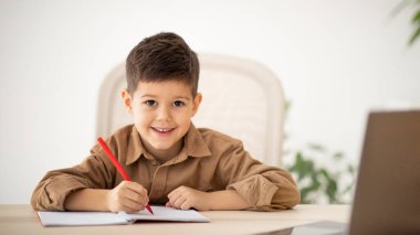 Video call, study remotely. Smiling caucasian little child sit at table, making notes, watching lesson on computer in room interior. Education at school, kindergarten and home, childhood and lifestyle