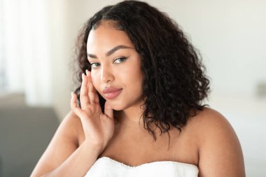 Beautiful black plus size woman touching face and looking at camera, body positive woman with flawless skin standing wrapped in towel, home interior