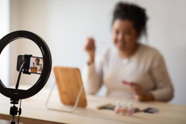 Selective focus on blogger set recording cute young plump hispanic woman beauty blogger sitting at table in front of mirror, recording video at home while applying lipstick, blurred background
