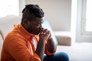 Pensive thoughtful sad young chubby black man in casual with dreadlocks sitting alone on couch at home, holding hands clenched together, touching his lips, looking down, copy space