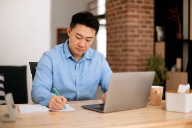 Serious asian middle aged man working with documents at home, sitting at desk with laptop and taking notes, checking financial reports, free space
