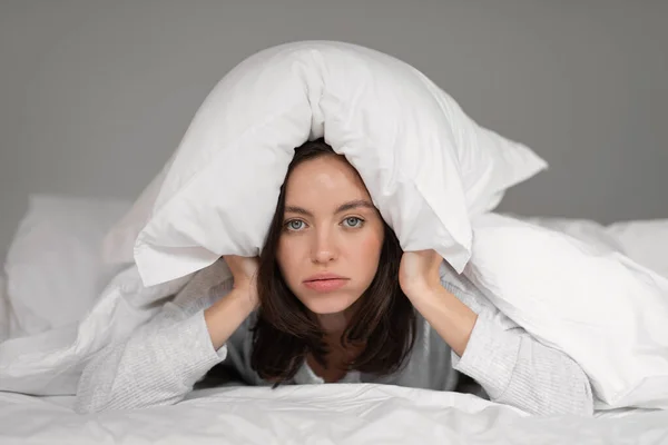 Unhappy tired young european woman lies on white comfortable bed under blanket, covers her head with pillow, wakes up in bedroom. Problems with rest and relax at home, insomnia and noisy neighbors