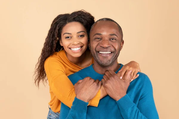 照片中 年轻快乐的黑人妇女从背后拥抱着她的中年丈夫 站在桃子工作室的背景上 随意的男人和女人看着相机笑着 — 图库照片