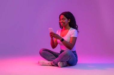 Smiling Young Black Woman Texting On Smartphone While Sitting On Floor In Neon Light Over Purple Studio Background, Cheerful African American Female Enjoying Online Communication, Copy Space