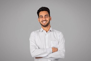 Smiling Handsome Young Arab Businessman With Folded Arms Standing Over Grey Background And Looking At Camera, Happy Middle Eastern Male Entrepreneur In White Shirt Posing In Studio, Copy Space