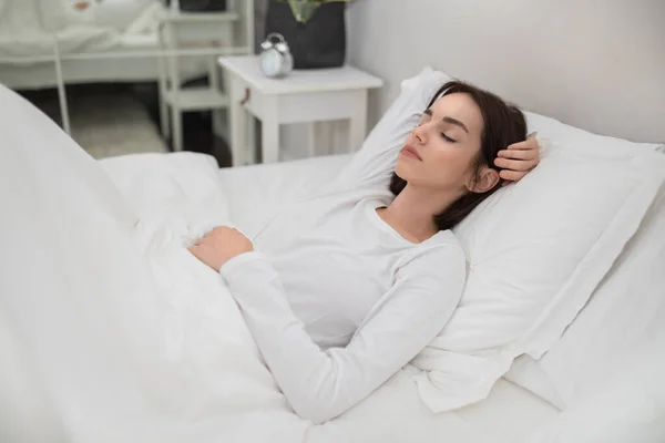 Pretty young brunette woman sleeping with hand under her head on soft comfy pillows, peaceful female lying in bed with closed eyes, candid photo. Resting, sleeping, relaxation at home concept