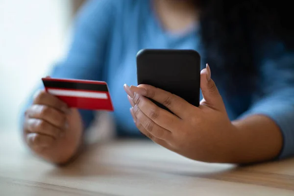 E-commerce, retail. Unrecognizable dark-skinned chubby woman in blue shirt sitting at desk indoors, holding modern smartphone and red bank card. Female customer banking or shopping online