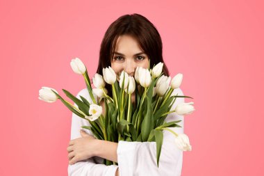 Cheerful young european lady smelling bouquet of flowers, enjoy lifestyle, aroma of tulips, rejoice spring festival isolated on pink background, studio. Romantic gift on date, birthday, ad and offer