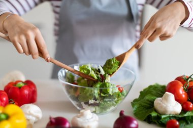 Unrecognizable woman wearing grey apron preparing healthy vegetable salad for dinner at home, female hands holding wooden spoons, mixing veggies in glass bowl, cropped. Diet, nutrition concept