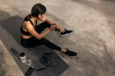 Tired slender caucasian millennial lady in sportswear sits on mat with skipping rope and bottle of water, checks pulse on street. Sports, fitness workout and active in city, body care and slimming