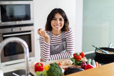 Cheerful positive beautiful young middle eastern woman with nice hairstyle cooking at home, using fresh organic vegetables, holding tomato in her hand and smiling at camera, copy space