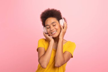 Cheerful young african american curly female in yellow t-shirt and wireless headphones with closed eyes enjoy music isolated on pink background, studio. Device to listen song, audio app in free time
