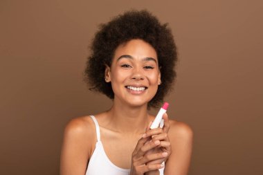 Cheerful young african american curly female with perfect skin in white top uses moisturizing lipstick for lips, isolated on brown background, studio, copy space. Beauty care, nude natural makeup