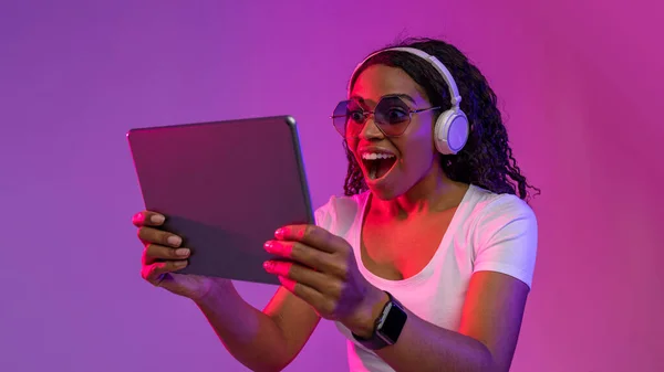 Portrait Of Amazed Black Female In Headphones And Stylish Eyeglasses Using Digital Tablet, Surprised African American Woman Listening Music Or Playing Video Games While Standing In Neon Light