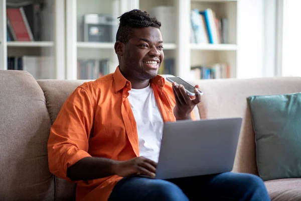 Happy cheerful african american guy businessman working from home, sitting on couch in living room, using modern laptop and smartphone, recording voice mail, looking at copy space and smiling