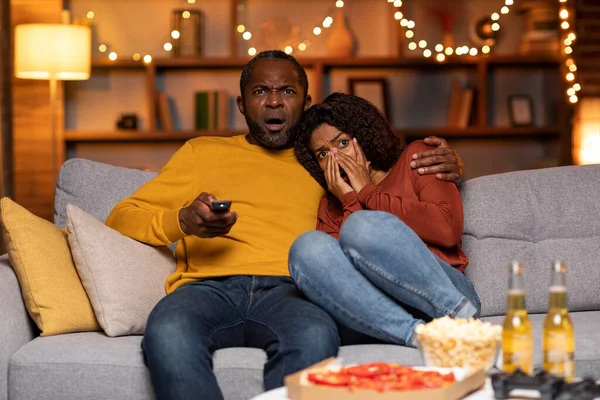 Frightened african american middle aged man and young woman watching horror movie on TV together at home, black couple sitting on sofa in cozy living room decorated with lights, copy space