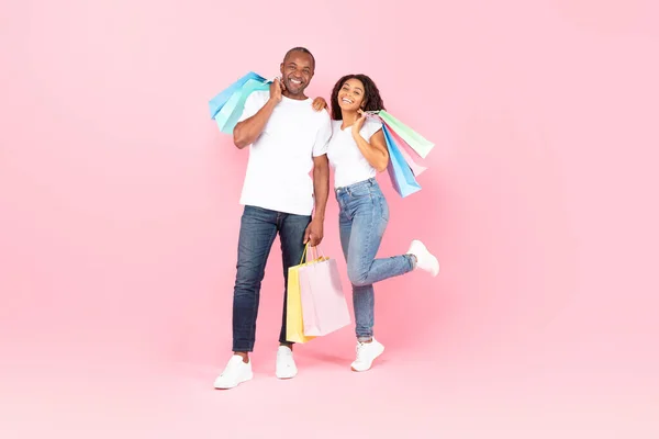 Happy african american couple with shopping bags posing on pink studio background, full length shot, free space. Positive black spouses buying gifts on sale, happy over big discounts