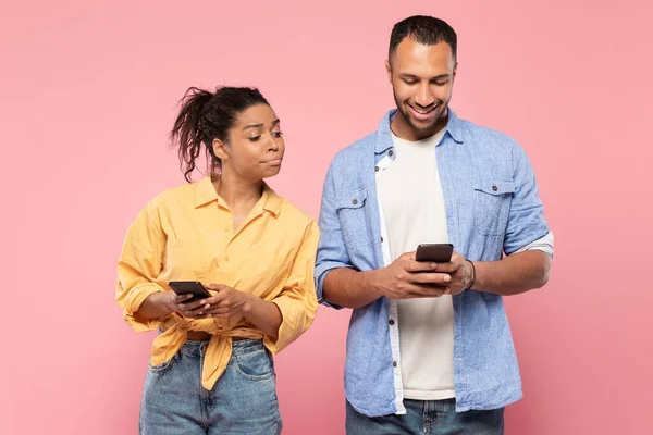 アフリカ系アメリカ人女性スパイオン彼女の笑顔ボーイフレンド誰を使用して携帯電話 テキストメッセージSmsまたはスクロールソーシャルメディアニュースフィード 一緒にピンクのスタジオの背景に立って — ストック写真