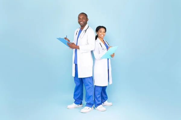 African american man and woman doctors in white coats with stethoscopes writing at tablet and smiling, blue background, full length. Medical service, examination with therapist, prescription
