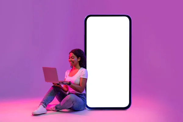 Smiling black lady with laptop leaning at big blank smartphone with white screen while sitting in neon light over purple studio background, happy female browsing new website, collage, mockup
