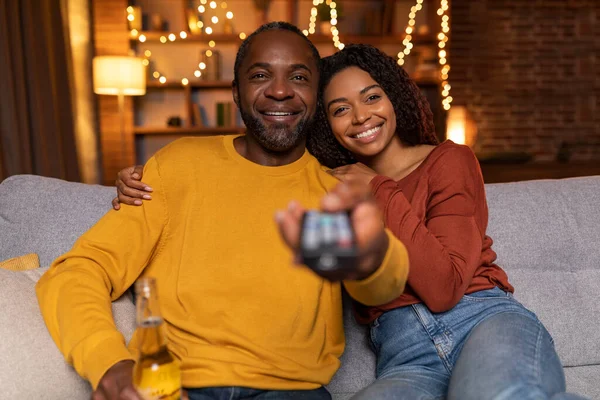 Family portrait of loving black couple enjoying romantic evening together at home, happy man and woman sitting on couch, cuddling, switching channels with remote control, watching TV, copy space