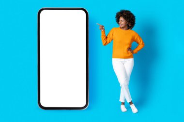 Cheerful smiling attractive curly millennial african american woman in stylish outfit point at huge cell phone with white blank screen, showing nice online offer, blue background, mockup, full length