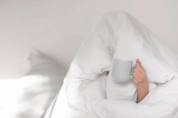 Sad tired funny caucasian millennial woman sitting on bed, covered with white blanket with coffee cup, dont wont awake in white bedroom interior, unrecognizable. Time to work, wake up early at home