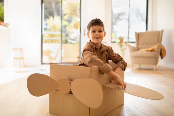 Cheerful cute caucasian little child sit in cardboard box airplane, has fun alone, enjoy travel in living room interior, sun flare. Entertainment and play game at home, fantasy, art and childhood