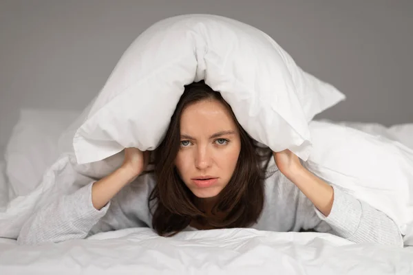 疲惫而悲伤的高加索千禧年女性躺在白色毯子下躺在床上 用枕头盖住头 在卧室里饱受失眠和噪音之苦 家里的压力 健康问题和抑郁症 — 图库照片