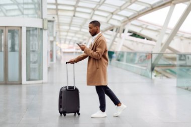 Online Check-In. Young Black Man Walking With Suitcase In Airport And Using Smartphone, African American Male Browsing Internet Or Messaging On Cellphone While Going To Flight Gate, Copy Space