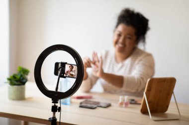 Cheerful pretty curly young chubby hispanic lady beauty blogger sitting at desk full of various makeup products, recording video on smartphone, showing different nail poloshers, recommending cosmetics